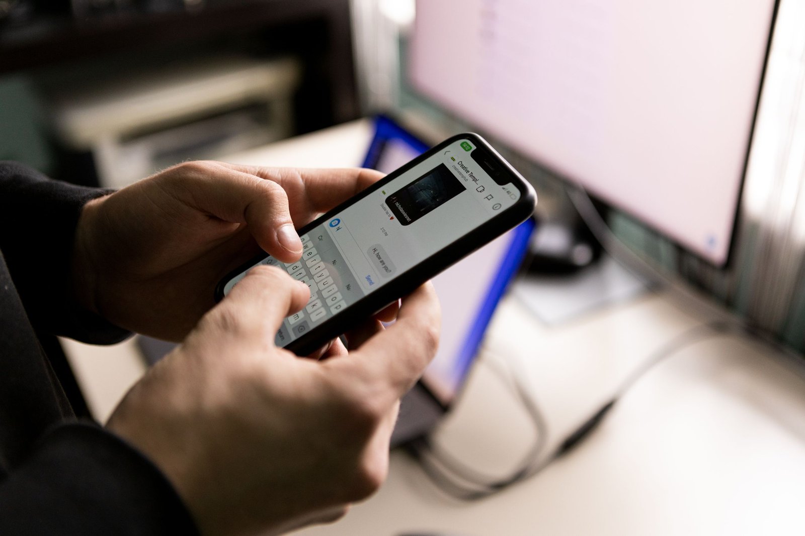 A person holding a smartphone and chatting indoors near a computer screen.